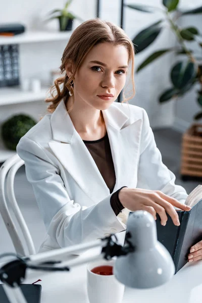 Hermosa mujer de negocios segura sentada en el escritorio, sosteniendo el cuaderno y mirando a la cámara en el lugar de trabajo - foto de stock