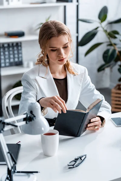Concentrated businesswoman in formal wear sitting at desk and reading notebook at workplace — Stock Photo