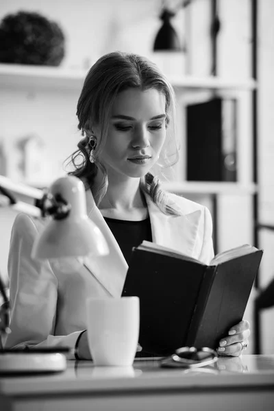 Black and white photo of businesswoman in formal wear sitting at desk and reading notebook at workplace — Stock Photo