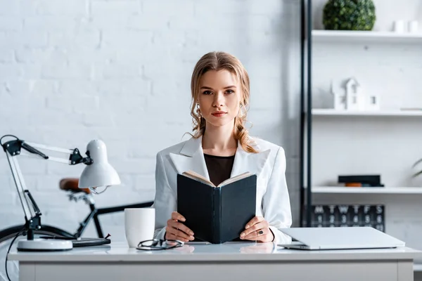 Empresaria concentrada sentada en el escritorio, sosteniendo el cuaderno y mirando la cámara en el lugar de trabajo - foto de stock