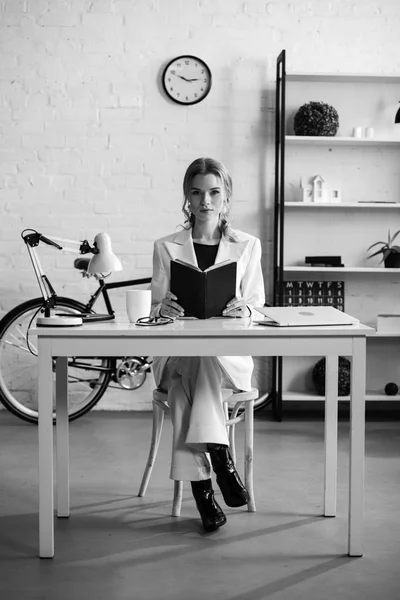 Negro y blanco de una mujer de negocios seria sentada en el escritorio, sosteniendo un cuaderno y mirando a la cámara en el lugar de trabajo - foto de stock