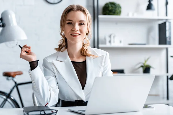 Lächelnde Geschäftsfrau in festlicher Kleidung sitzt am Schreibtisch, hält Stift in der Hand und blickt am Arbeitsplatz in die Kamera — Stockfoto