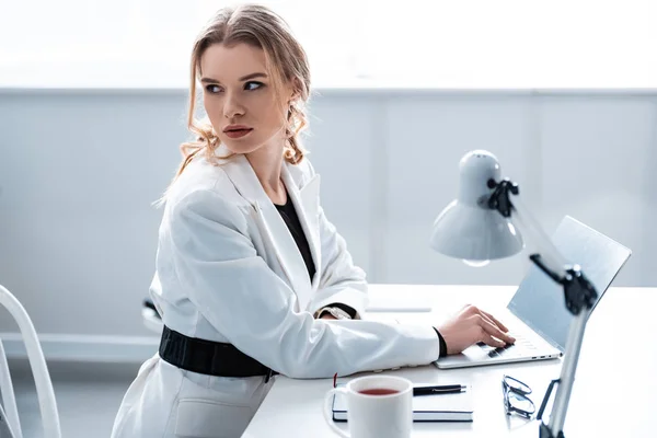 Bela mulher de negócios em desgaste formal sentado na mesa e olhando para longe ao usar laptop no local de trabalho — Fotografia de Stock