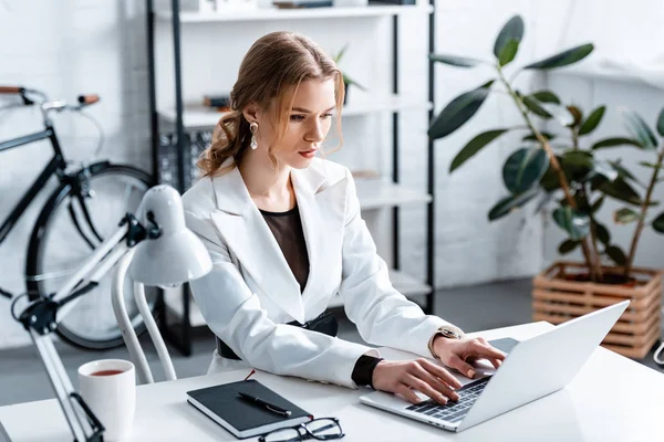 Belle femme d'affaires ciblée en tenue formelle assis au bureau et tapant sur ordinateur portable sur le lieu de travail — Photo de stock