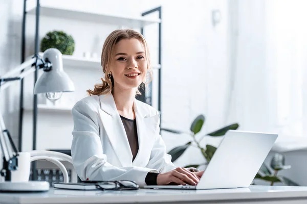Femme d'affaires souriante en tenue de cérémonie assise au bureau, regardant la caméra et tapant sur ordinateur portable sur le lieu de travail — Photo de stock