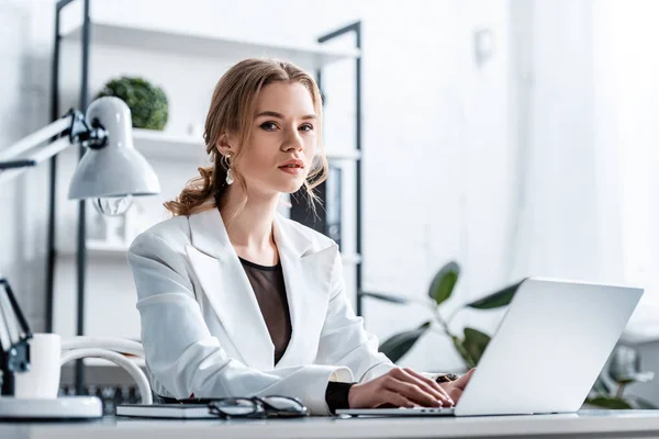 Mujer de negocios seria en ropa formal sentado en el escritorio, mirando a la cámara y escribiendo en el ordenador portátil en el lugar de trabajo - foto de stock