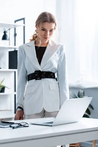 Mujer de negocios seria en ropa formal de pie cerca de escritorio y mirando a la computadora portátil en la oficina - foto de stock
