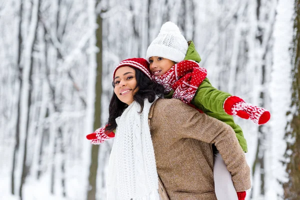 Attractive african american woman giving piggyback ride to smiling daughter in winter park — Stock Photo