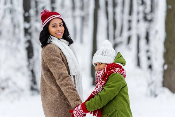 Sorridente afro-americana mãe e pré-adolescente filha de mãos dadas e olhando para a câmera no parque de inverno — Fotografia de Stock