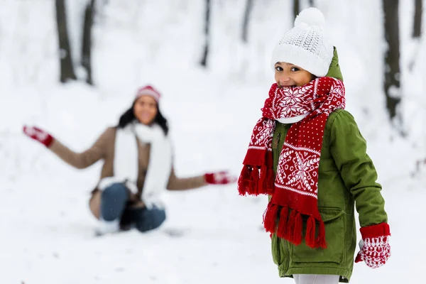 Bonito criança olhando para câmera e afro-americana mãe no fundo no parque de inverno — Fotografia de Stock