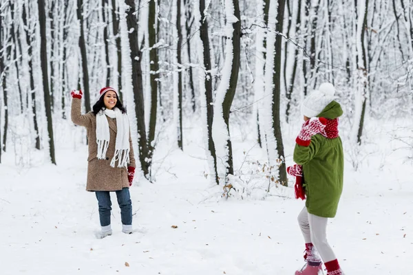 Fröhliche afrikanisch-amerikanische Mutter lächelt und spielt Schneebälle mit Tochter in warmer Kleidung im Winterpark — Stockfoto