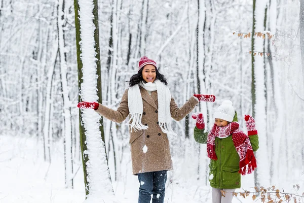 Cheerful african american woman and preteen daughter throwing up snow in winter park — Stock Photo