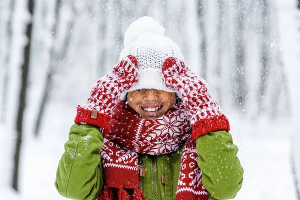 Niño afroamericano sonriente con sombrero de punto tirado sobre los ojos durante las nevadas en el parque de invierno - foto de stock