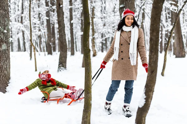 Beautiful african american woman smiling and pulling preteen daughter in warm clothing on sledge in winter park — Stock Photo