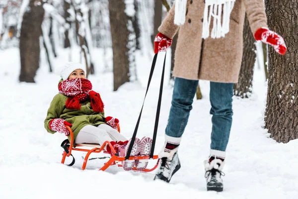 Abgeschnittene Ansicht von Mutter, die Tochter in warmer Kleidung auf Schlitten im Winterpark zieht — Stockfoto