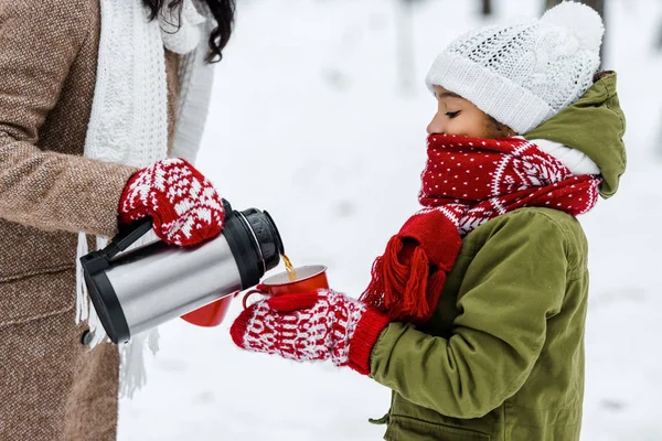 Vista ritagliata della madre versando il tè dal thermos in tazza rossa tenuta dalla figlia afro-americana in inverno — Foto stock