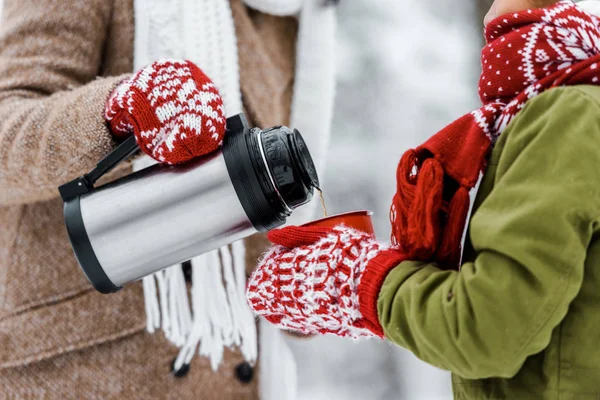 Vue recadrée de la femme versant le thé du thermos dans la tasse rouge tenant par l'enfant en hiver — Photo de stock