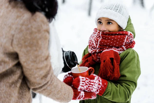 Cropped view of mother giving tea to cute african american daughter in snowy forest — Stock Photo