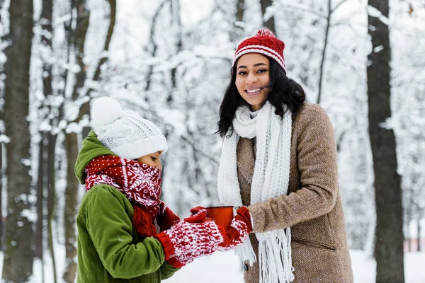 Atractiva madre afroamericana y linda hija sosteniendo tazas de té y sonriendo a la cámara en el bosque nevado - foto de stock