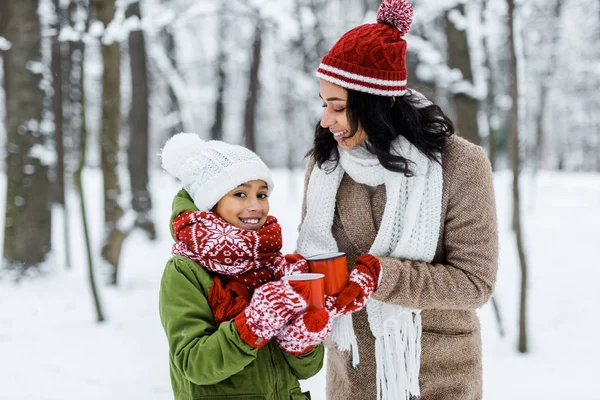 Atractiva madre afroamericana y linda hija sosteniendo tazas de té y mirando a la cámara en el bosque nevado - foto de stock