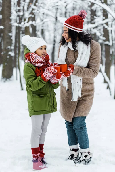 Atractiva madre afroamericana y linda hija hablando entre sí y sosteniendo tazas de té en el bosque nevado - foto de stock