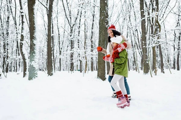 Attractive african american mother and cute daughter walking and holding cups in snowy forest — Stock Photo