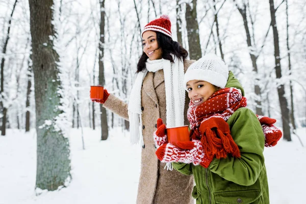 Atractiva madre afroamericana y linda hija caminando y sosteniendo copas rojas en el bosque nevado - foto de stock