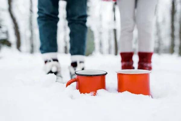 Vista recortada de tazas rojas de té que permanecen en la nieve en el parque en el bosque nevado - foto de stock