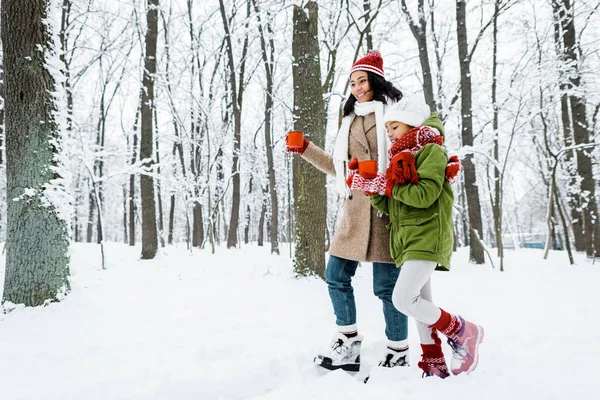 Attraente afro americano madre e carina figlia a piedi e in possesso di coppe rosse tra gli alberi e guardando la neve nella foresta innevata — Foto stock