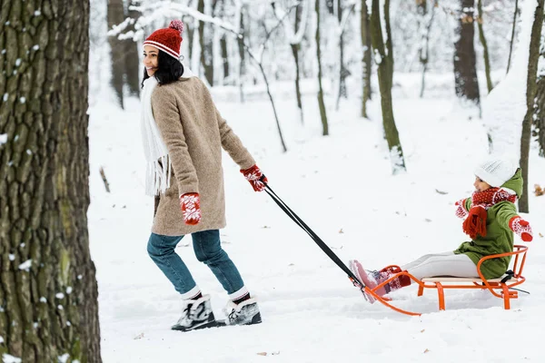 Afro-americana filha com mãe equitação no trenó perto de árvores e sorrindo na floresta nevada — Fotografia de Stock