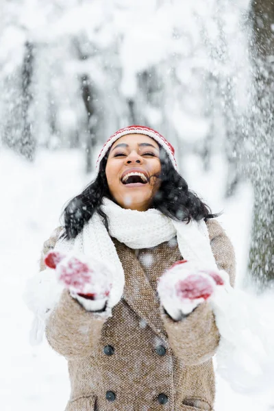 Atractiva mujer afroamericana lanzando nieve y sonriendo en el bosque de invierno - foto de stock