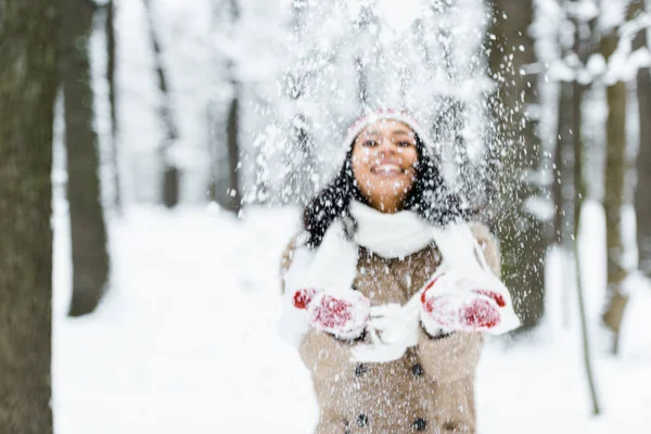 Привлекательная американка из Африки бросает снег в парк и улыбается в зимнем лесу — стоковое фото