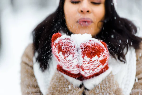 Cropped view of beautiful african american woman blowing snow in winter forest and looking at snow — Stock Photo