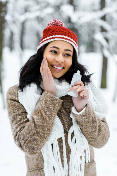Atractiva mujer afroamericana tocando mejilla y sonriendo en el bosque de invierno - foto de stock