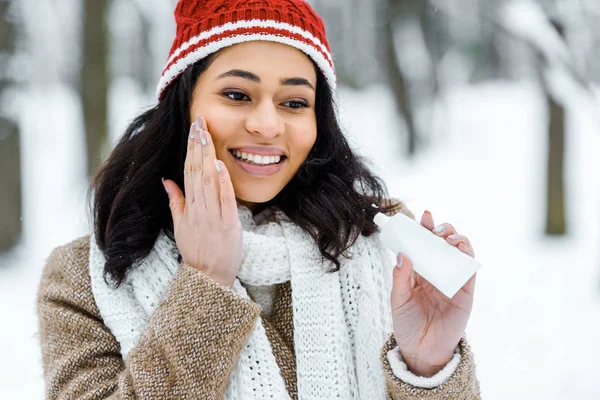 Atractiva mujer afroamericana tocando mejilla y aplicando crema protectora en el bosque de invierno - foto de stock
