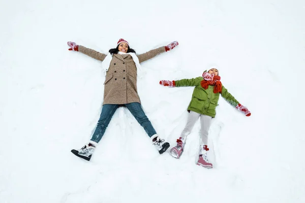 Attraente afro americano madre e carino figlia fare neve angeli in inverno foresta — Foto stock