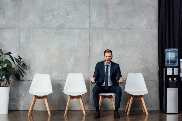 Hombre de negocios sentado en la silla y animando con los puños cerrados en la sala de espera - foto de stock