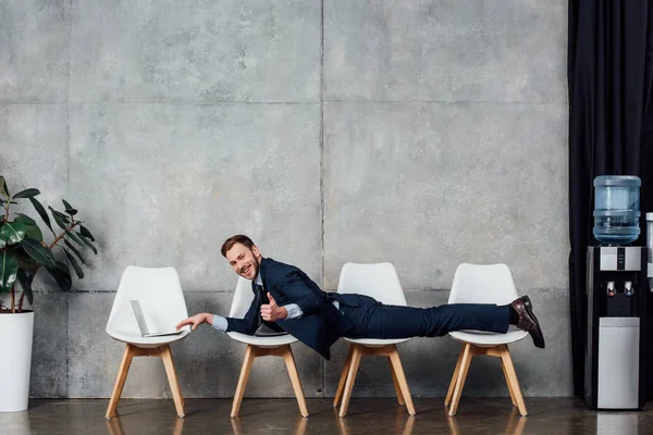 Businessman lying on chairs, showing thumb up sign and using laptop in waiting hall — Stock Photo