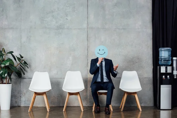 Businessman in suit sitting on chair and holding card with winking face expression in waiting hall — Stock Photo