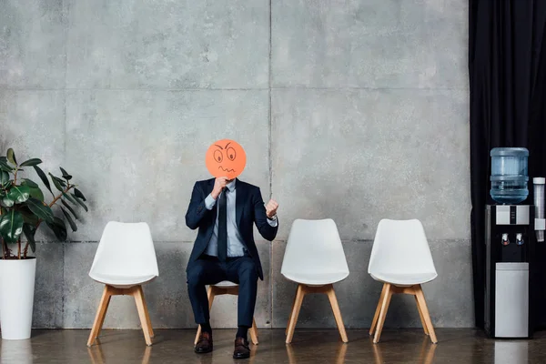 Бизнесмен в костюме сидит на стуле и держит карточку с запутанным лицом в зале ожидания — стоковое фото