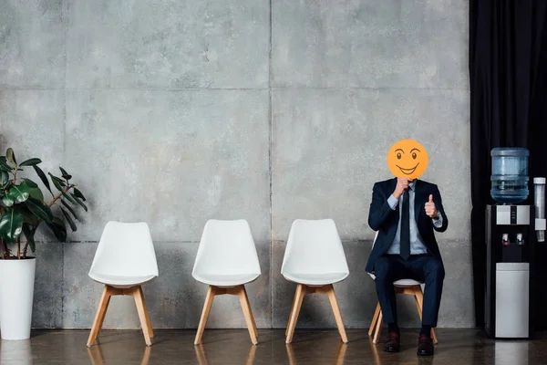 Uomo d'affari in giacca e cravatta seduto e in possesso di carta con espressione faccia sorridente in sala d'attesa e mostrando pollice su segno — Foto stock