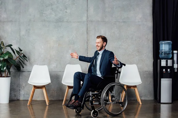Hombre de negocios sonriente con las manos extendidas sentado en silla de ruedas en la sala de espera - foto de stock