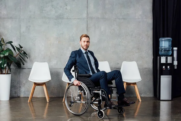 Hombre de negocios serio sentado en silla de ruedas en la sala de espera y mirando a la cámara - foto de stock