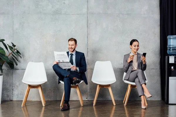 Empresario leyendo periódico mientras asiático mujer de negocios usando smartphone en sala de espera - foto de stock