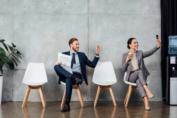 Hombre de negocios con periódico saludando con la mano mientras que la mujer de negocios asiática tomando selfie en el teléfono inteligente en la sala de espera - foto de stock