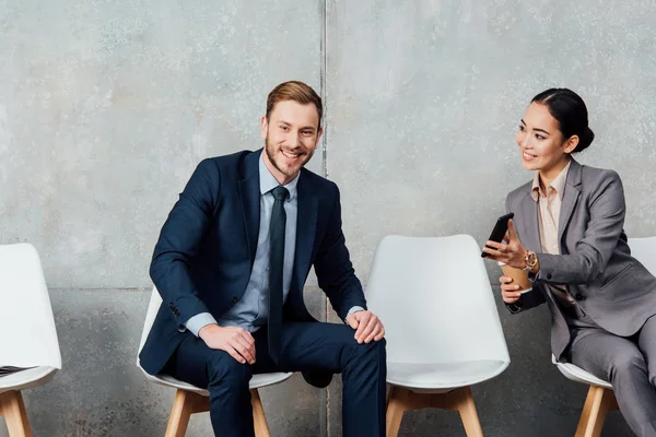 Handsome smiling businessman looking at camera while asian businesswoman holding smartphone in waiting hall — Stock Photo