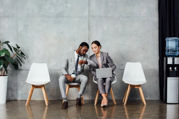 Multiethnic colleagues with laptop and coffee sitting and checking time on watch in waiting hall — Stock Photo