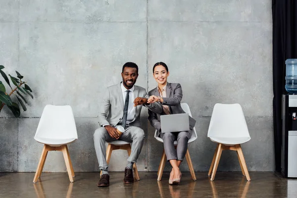 Sonrientes colegas multiétnicos con portátil y café sentado y comprobando el tiempo en los relojes en la sala de espera - foto de stock