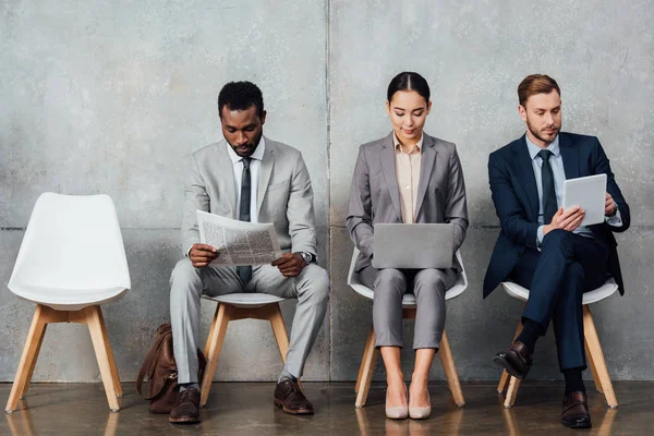 Empresarios multiétnicos enfocados en leer periódicos y usar dispositivos digitales en la sala de espera - foto de stock