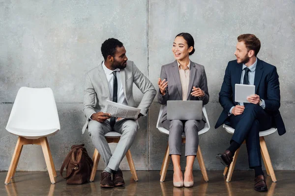 Multiethnic businesspeople reading newspaper, using digital devices and talking in waiting hall — Stock Photo